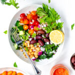 A salad bowl with kalamata olives, farro, chickpeas, tomatoes, cucumber, garnished with parsley and a roasted red pepper sauce.