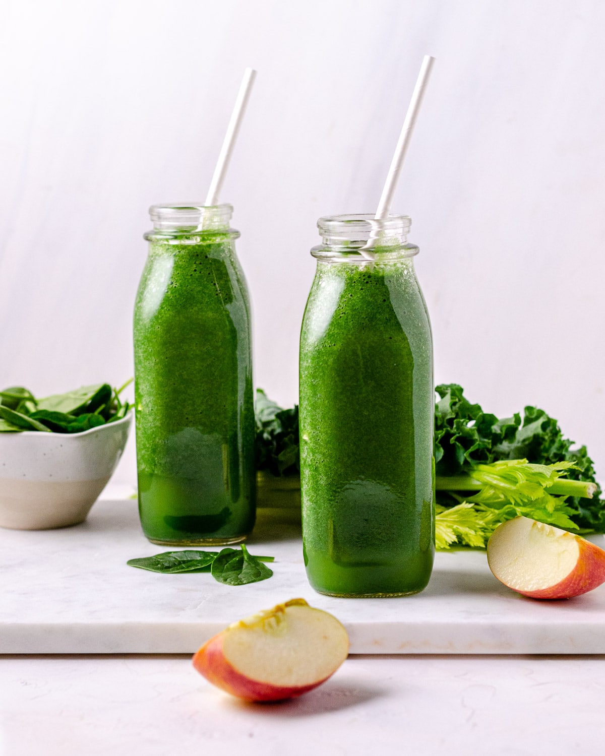 Two bottles of green smoothies on a cutting board with apples kale and celery,