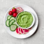 A bowl of vegan avocado green goddess dressing on a plate with rounds of cucumber, daikon radish and radishes