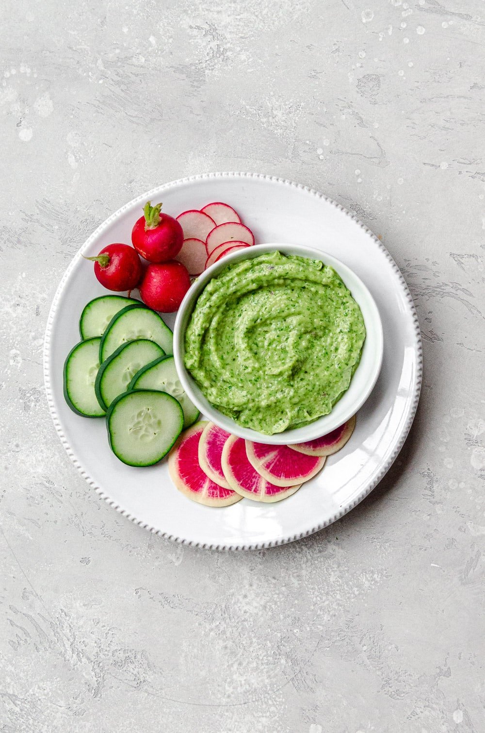 A bowl of vegan avocado green goddess dressing on a plate with rounds of cucumber, daikon radish and radishes