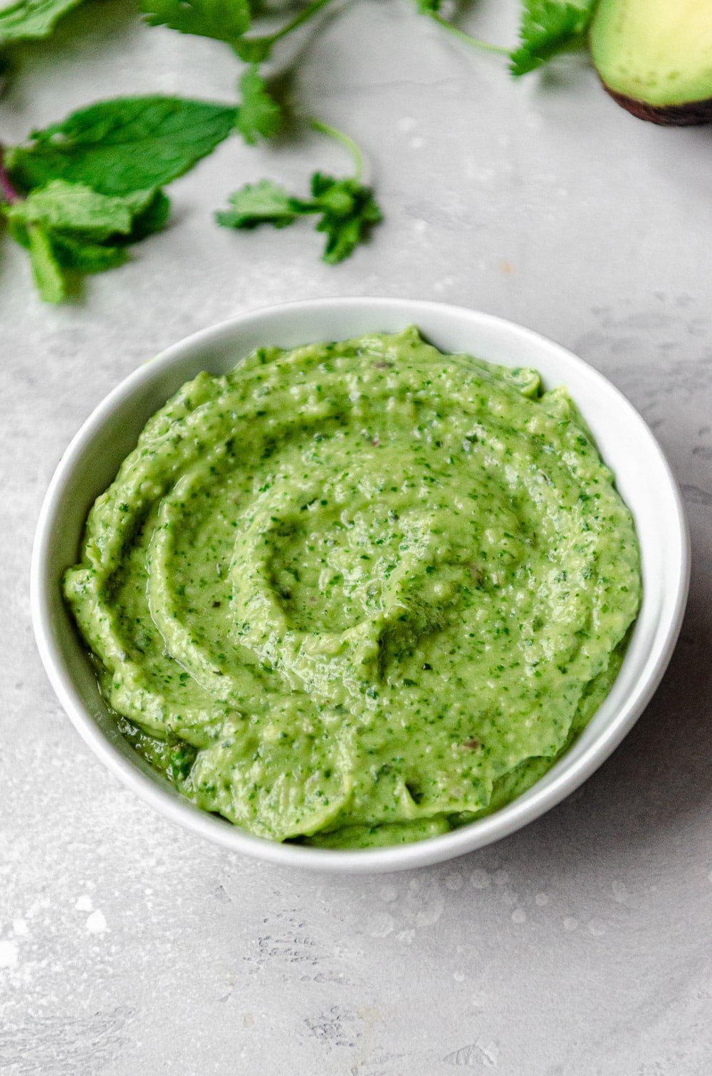 Close up of vegan green goddess dressing which is made from avocados. Surrounded by herbs and an avocado.