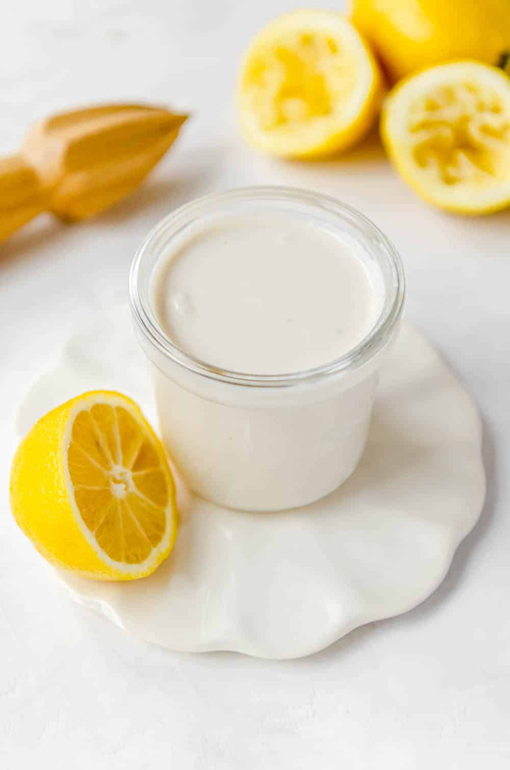 Close-up photo of jar of oil-free tahini dressing on a small coaster surrounded by lemons. 