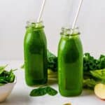 Two bottles of green blender smoothie with a cup of baby spinach, kale, celery and apples