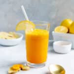 Glass of a turmeric ginger smoothie with lemon and coconut water, with a straw, next to a bowl of lemons and two small bowls of sliced turmeric and ginger.