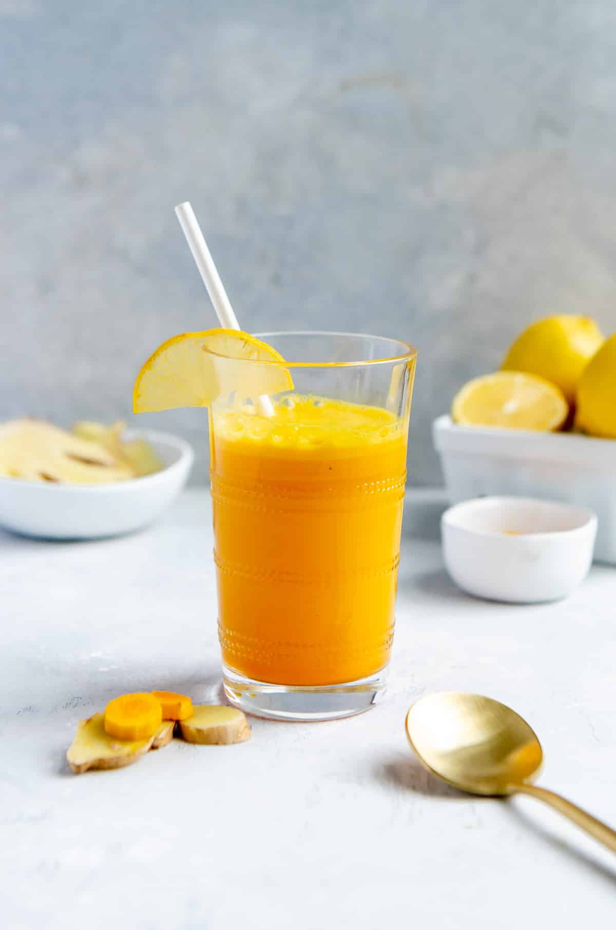 Glass of a turmeric ginger smoothie with lemon and coconut water, with a straw, next to a bowl of lemons and two small bowls of sliced turmeric and ginger.