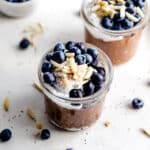 Healthy chocolate overnight oats with almond butter in two small jars garnished with coconut yogurt, almond slivers, chia seeds and blueberries.