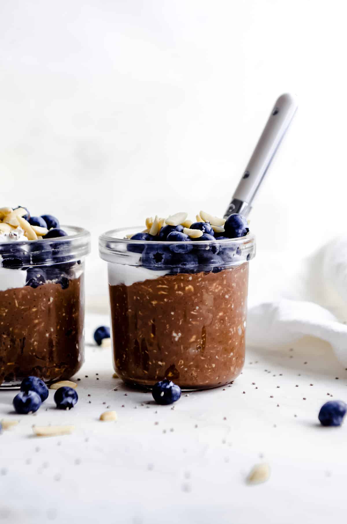 Small jar chocolate overnight oats with almond butter with coconut yogurt, almond slices and blueberry garnish, with a spoon sticking out of the jar.