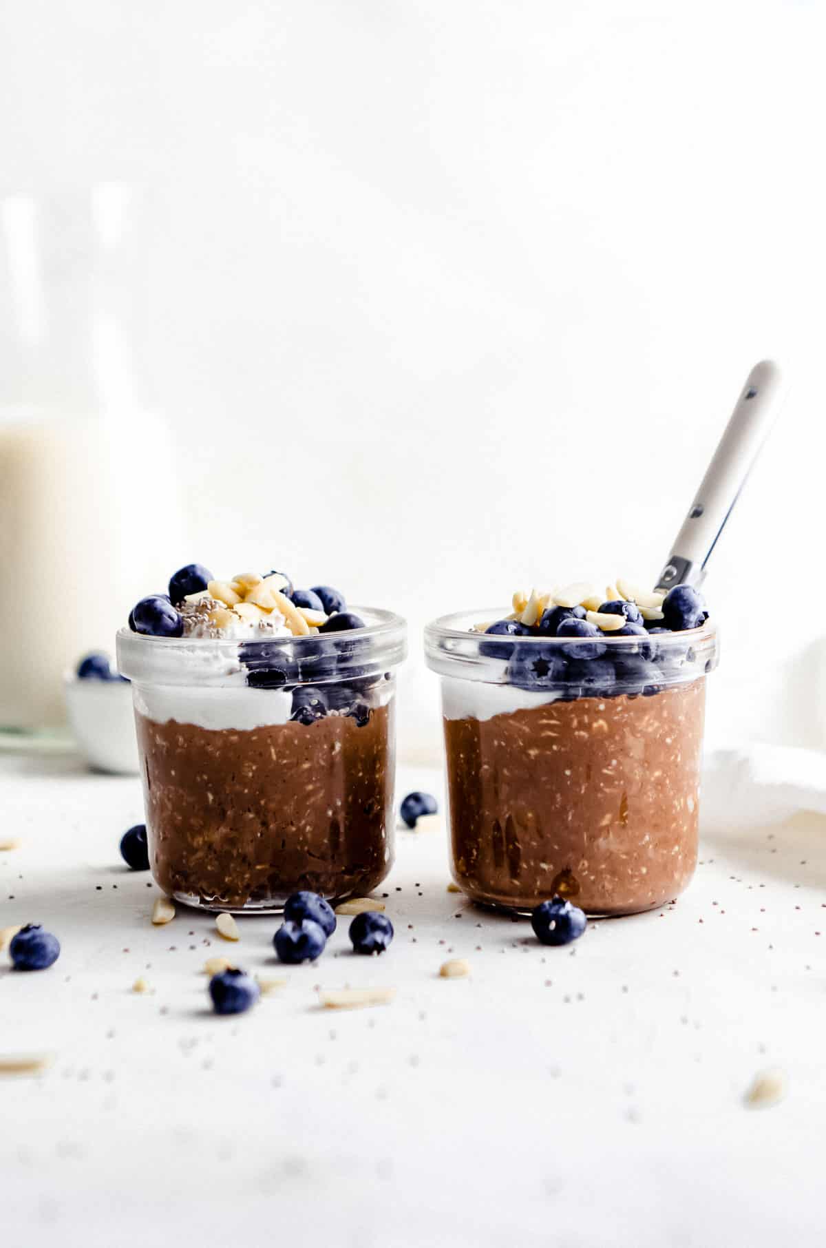 Two jars side by side of chocolate overnight oats garnished with coconut yogurt, almond slivers, blueberries and chia seeds.