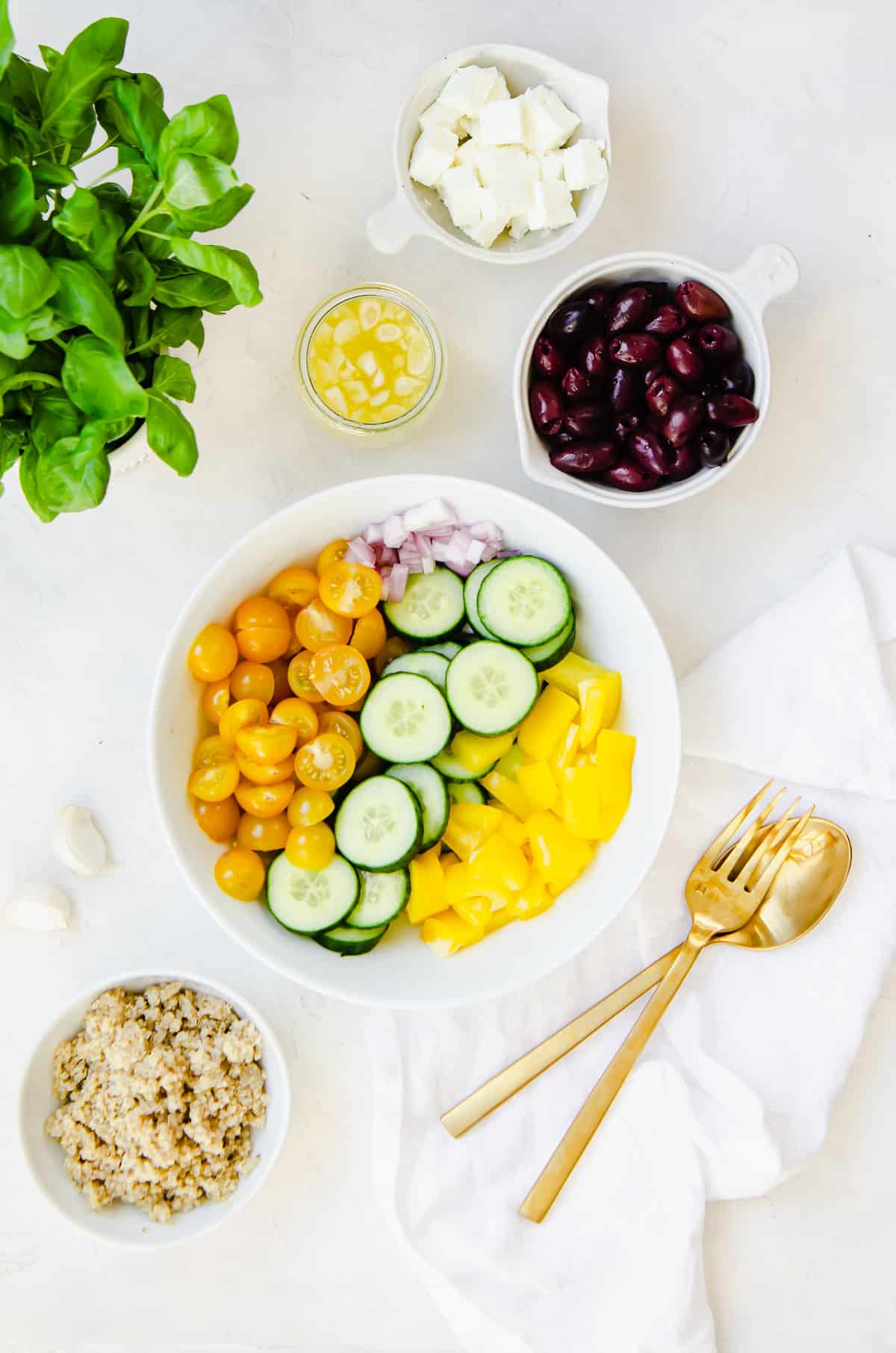 Ingredients for Mediterranean cucumber tomato salad in a bowl with a side of quinoa, basil leaves, garlic cloves, olives and a lemon dressing.