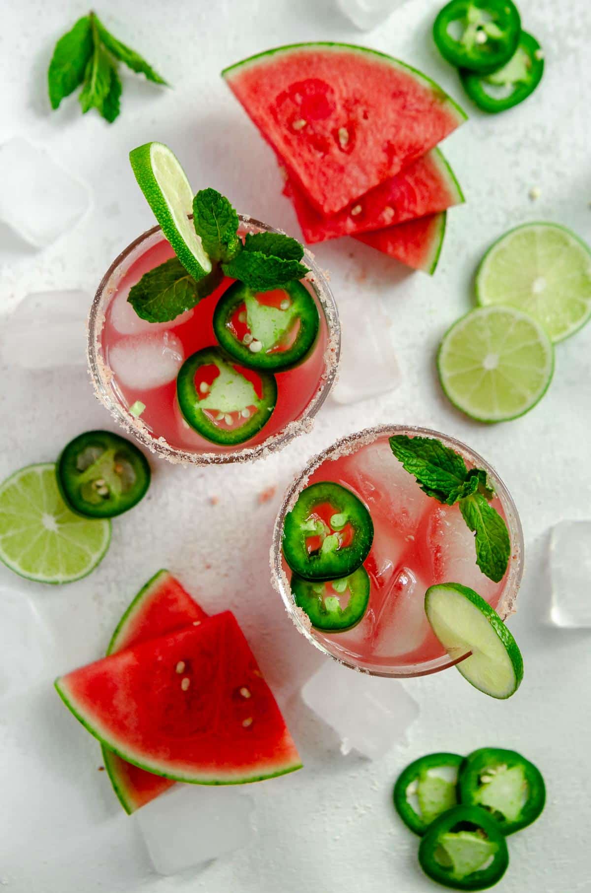 Two glassed of spicy watermelon margarita with pink salt rim and garnished with jalapeño, mint and watermelon slices