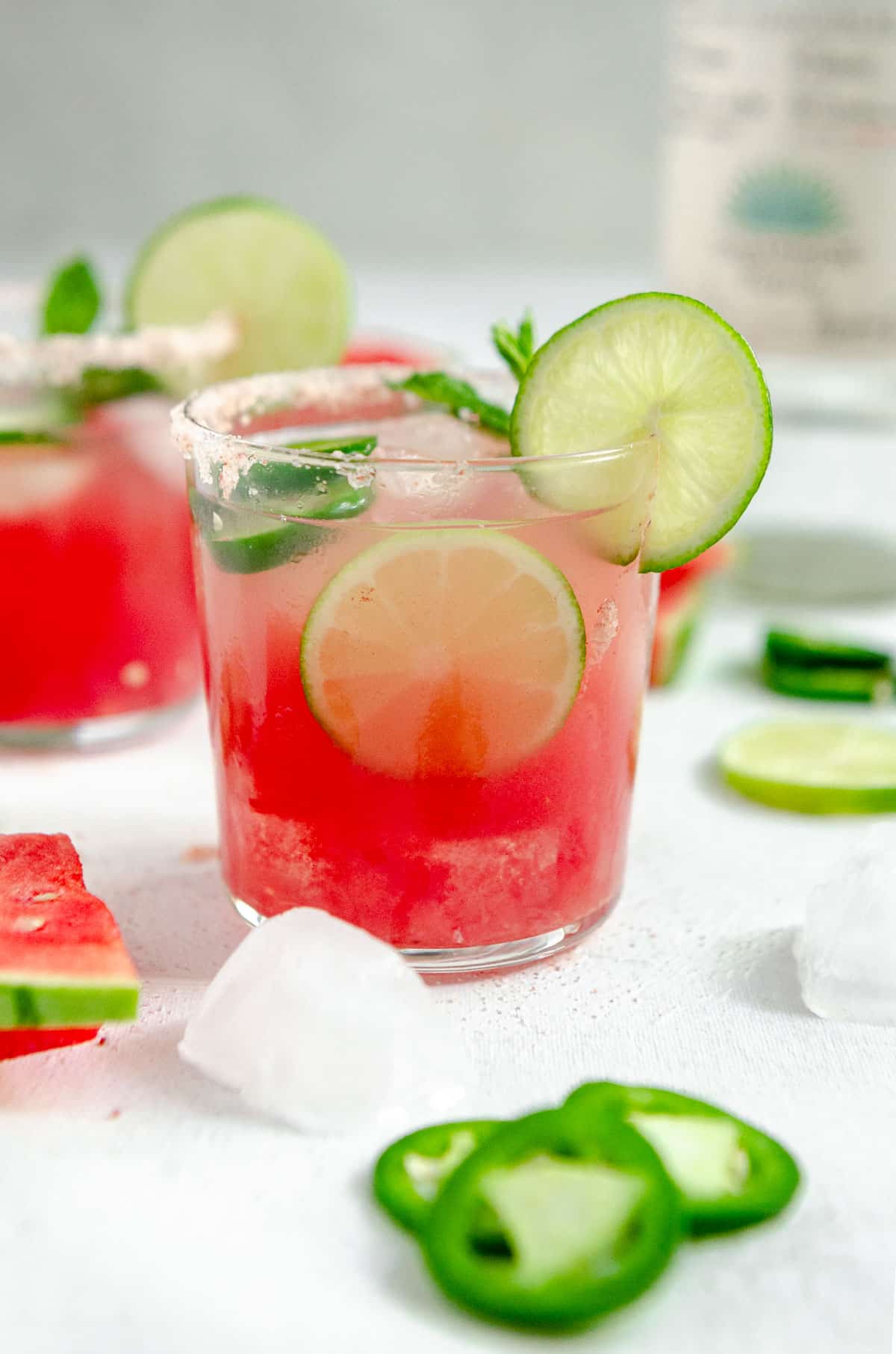 Glass of spicy watermelon margarita with pink salt rim and garnished with jalapeño, mint and watermelon slices with a bottle of Casamigos Blanco