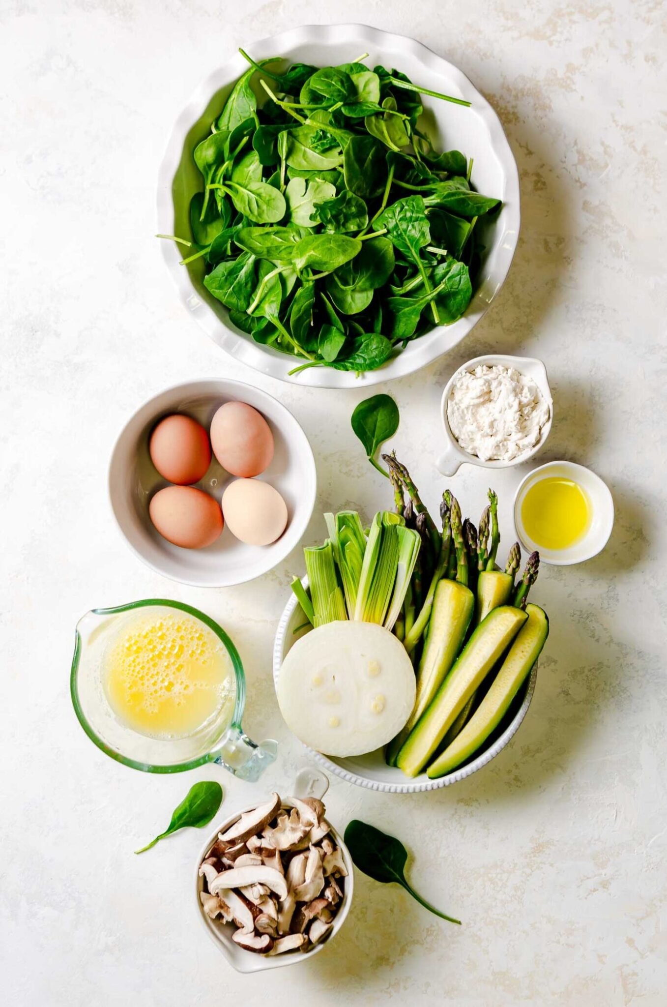 Ingredients for a green vegetable frittata including baby spinach, eggs, zucchini, onion, leeks, asparagus, ricotta, shiitake mushrooms.