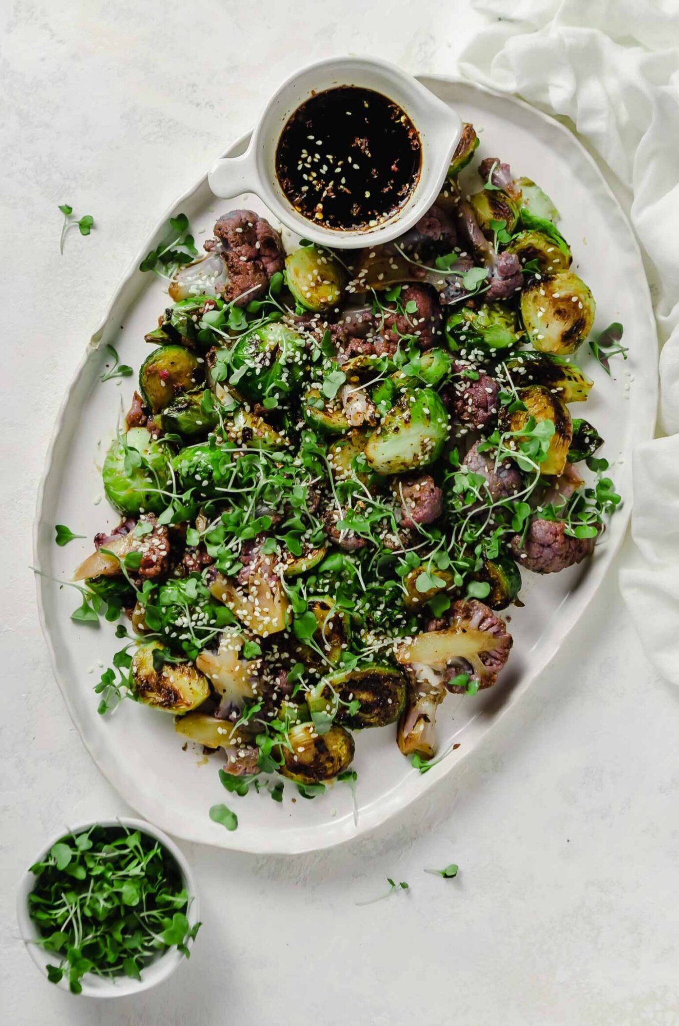 Plate of honey soy brussels sprouts garnished with sesame seeds, broccoli sprouts with a small bowl of extra sauce for dipping.