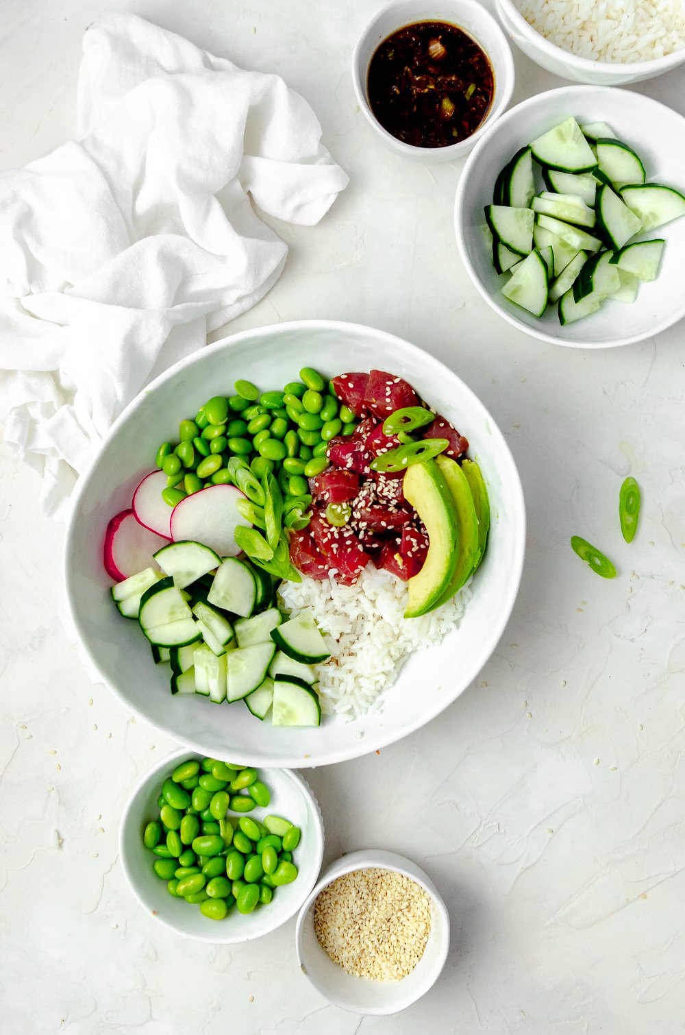 Tuna poke bowl made with edamame, white rice, radish, cucumbers and avocado, topped with sesame sesame and scallions. Additional bowls of ingredients surround the poke bowl. 