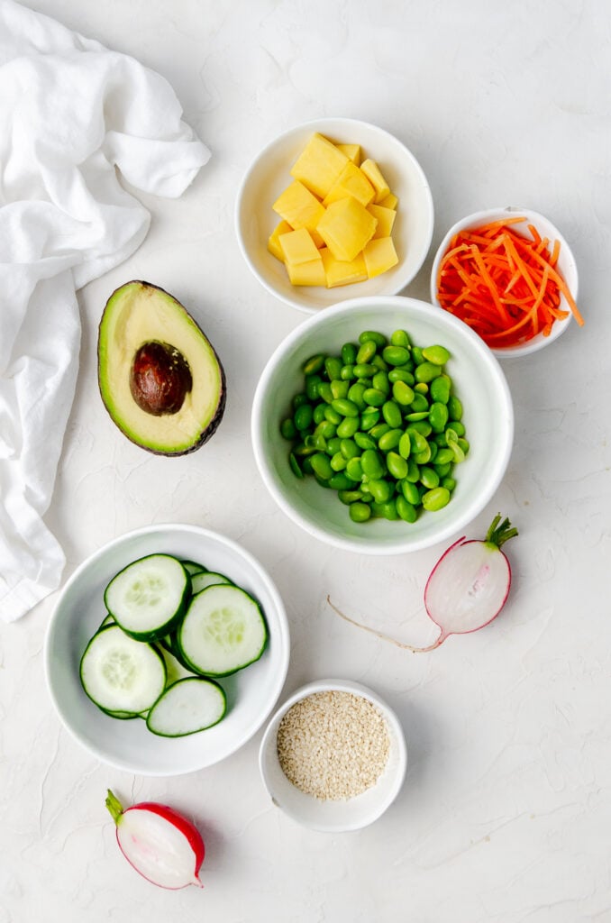 Potential toppings for a poke bowl which include mango, shredded carrots, avocado, edamame, sliced cucumbers or radish. 