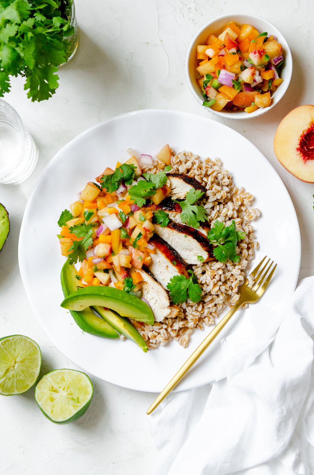 Plate of grilled chicken over farro, topped with a peach salsa made from peaches, yellow bell pepper, red onion, jalapeño and cilantro. Dish is surrounded by ingredients such as cilantro, limes and the salsa.