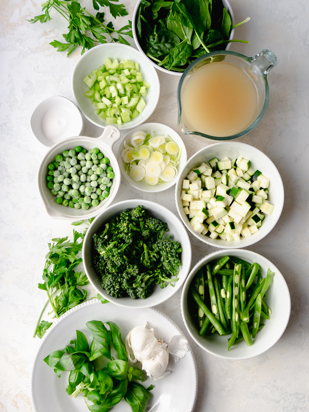 Green minestrone ingredients arranged in bowls - baby spinach, leeks, garlic, celery, frozen peas, broccolini, stringbeans, chicken broth, garlic, basil and parsley