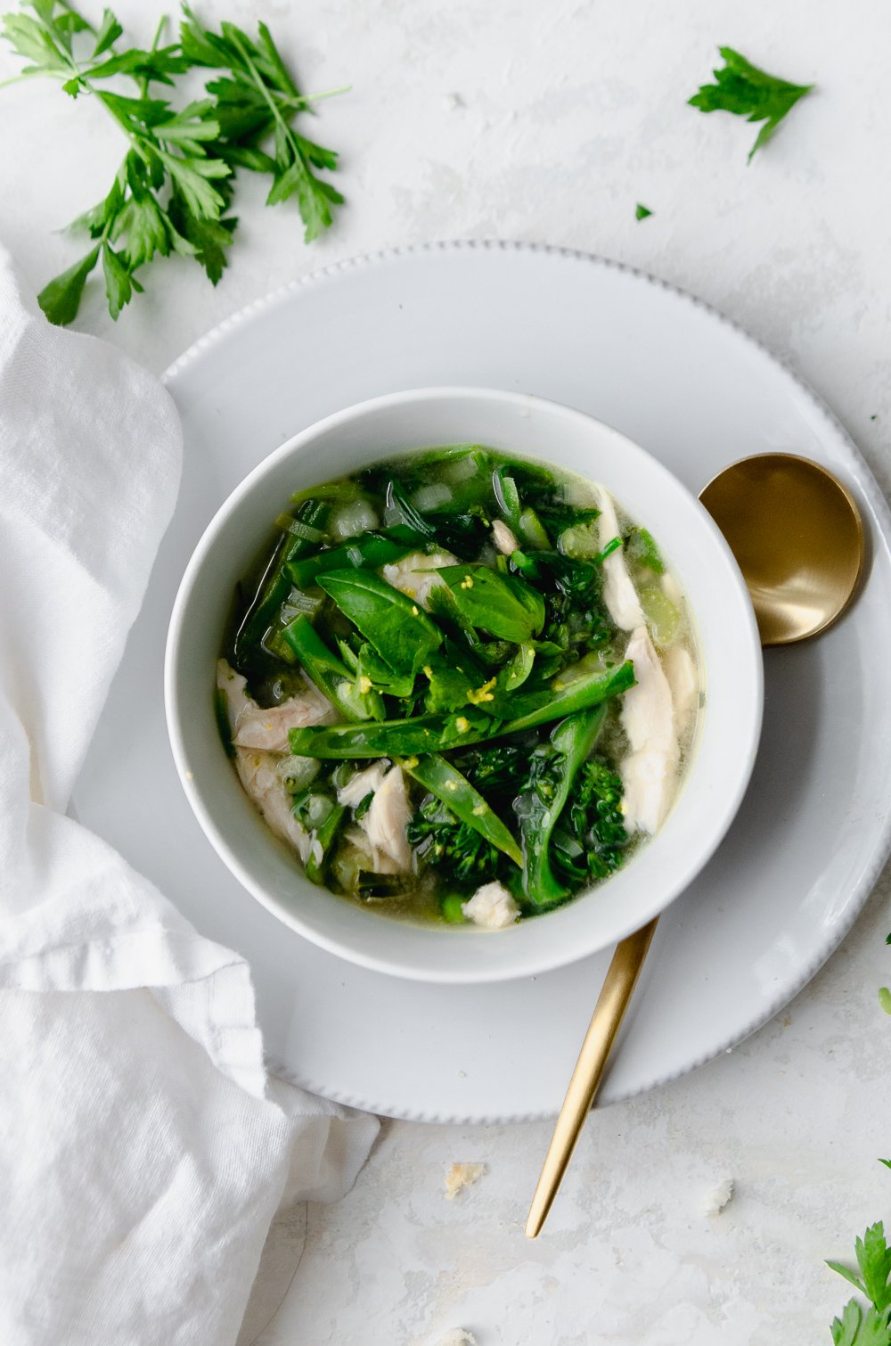 A white bowl of green vegetable minestrone soup with chicken on a white saucer with a gold spoon. 