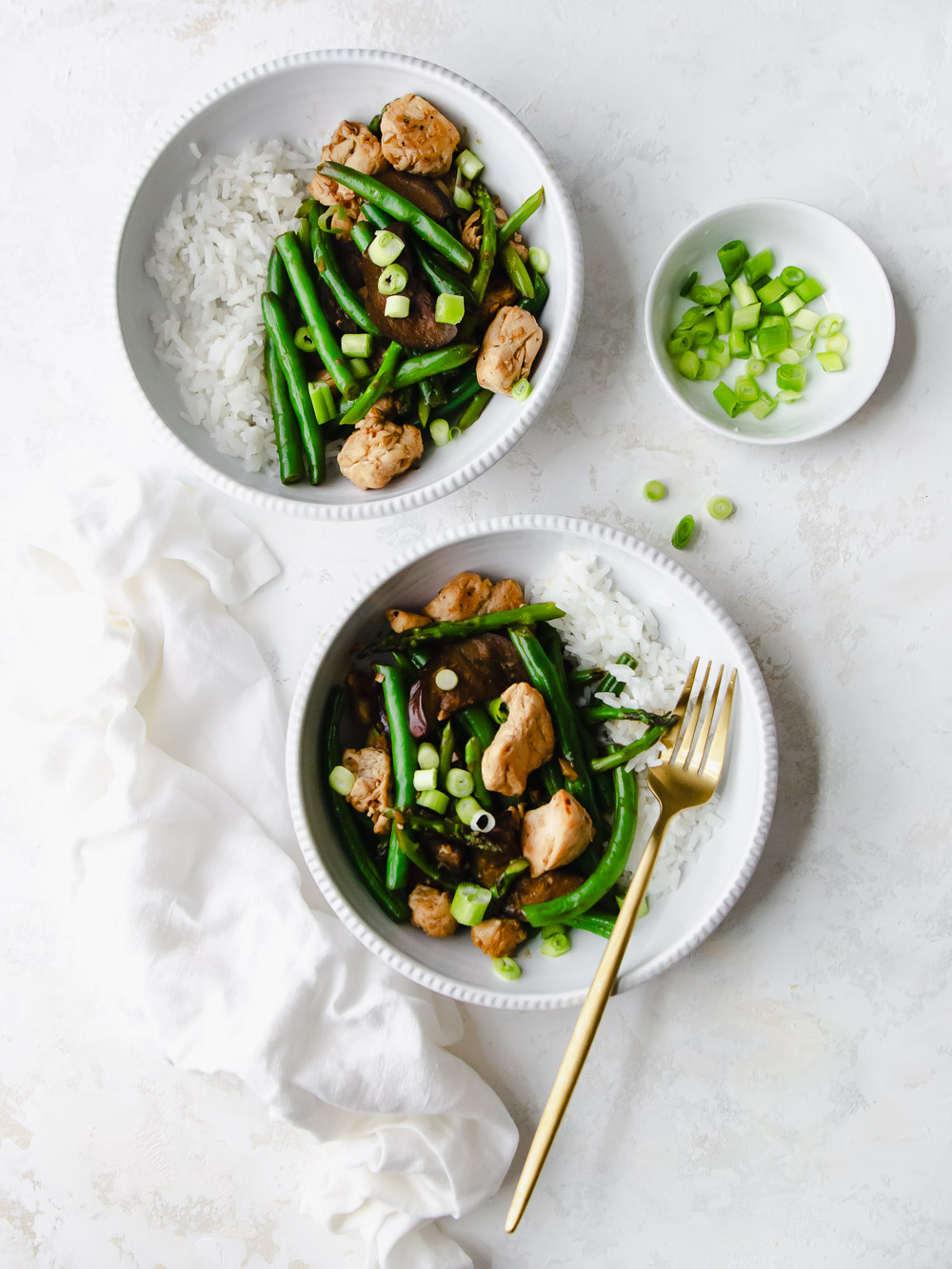 Two white bowls of healthy chicken stir fry made with green beans, egg plant and asparagus mixed with a garlic ginger (no soy) stir fry sauce, plated with white rice and garnished with green onions.