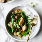 Close up of a white bowl of healthy chicken stir fry made with green beans, egg plant and asparagus mixed with a garlic ginger (no soy) stir fry sauce, plated with white rice, garnished with green onions and placed with a white napkin.