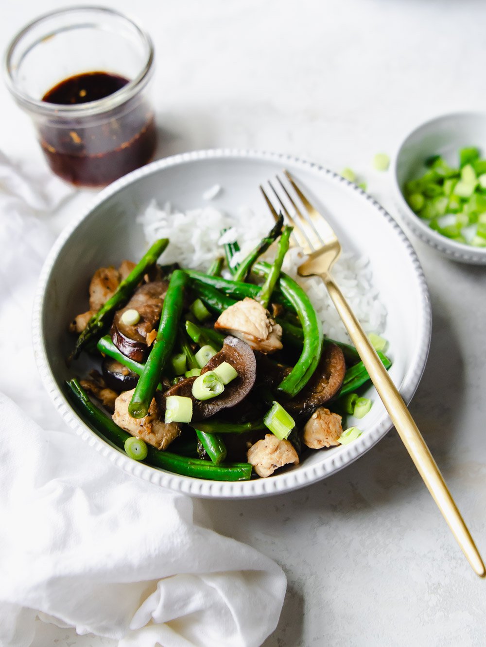 Bowl of chicken vegetable stir fry with string beans, eggplant and asparagus. Garnished with green onions and a gold fork.
