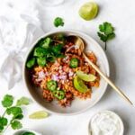 Mexican chicken and cauliflower rice recipe in a white bowl garnished with jalapeños, limes red onion and cilantro. With a gold serving spoon and surrounded by additional garnishes.