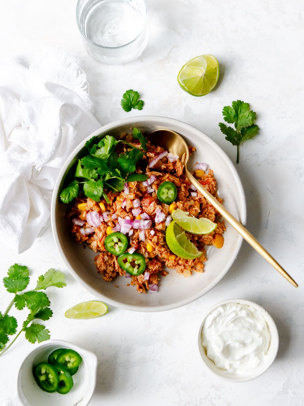 Mexican chicken and cauliflower rice recipe in a white bowl garnished with jalapeños, limes red onion and cilantro. With a gold serving spoon and surrounded by additional garnishes.