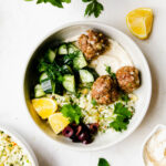 Three baked Greek meatballs nestled into herbed cauliflower rice. Dish is garnished with olives, lemons, cucumber salad, parsley and mint.