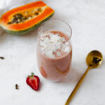 Papaya and strawberry smoothie topped with coconut flakes placed next to a gold spoon, strawberry and half of a papaya.