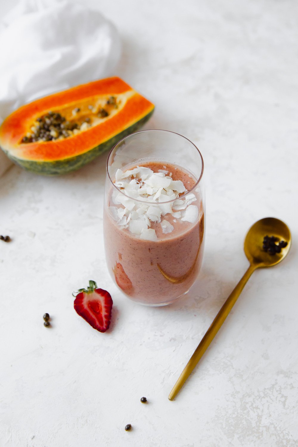 Papaya and strawberry smoothie topped with coconut flakes placed next to a gold spoon, strawberry and half of a papaya.