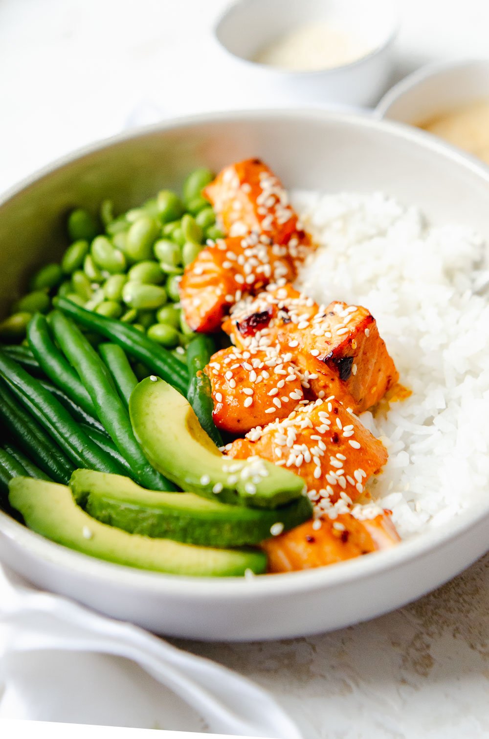 Hot honey salmon bites in a white bowl with white rice, avocado slices, string beans, edamame and is garnished with sesame seeds.