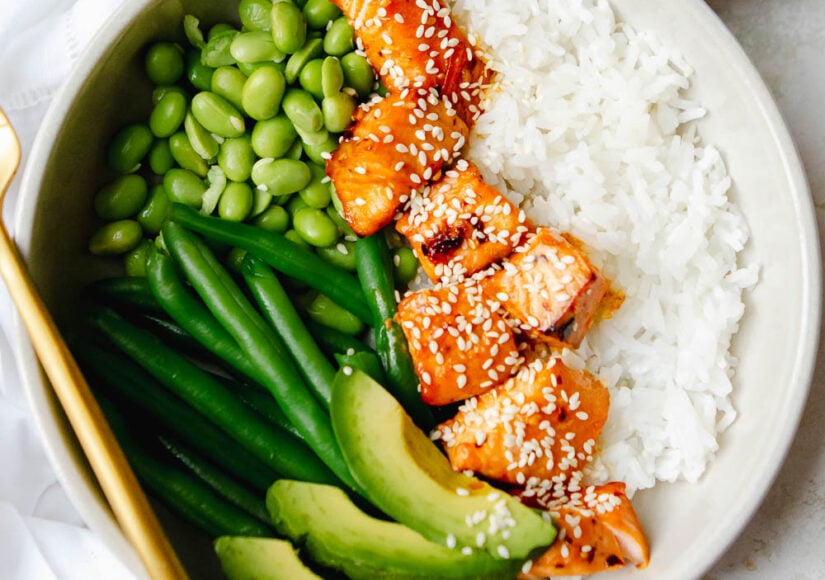 Close up of Hot honey salmon bites in a white bowl with white rice, avocado slices, string beans, edamame and is garnished with sesame seeds.