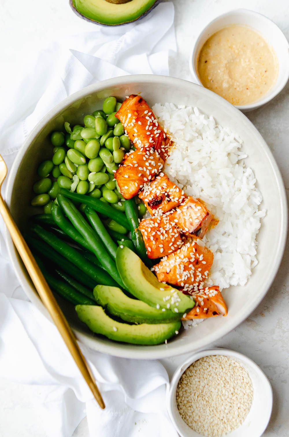 Close up of Hot honey salmon bites in a white bowl with white rice, avocado slices, string beans, edamame and is garnished with sesame seeds.