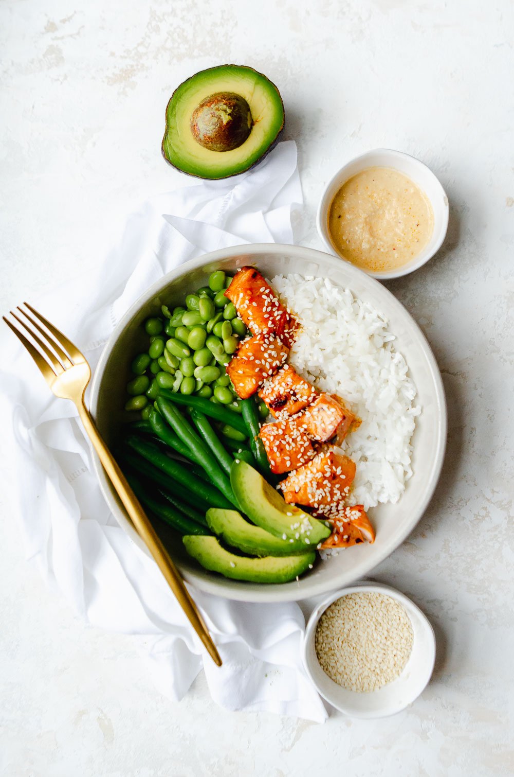 Hot honey salmon bites in a white bowl with white rice, avocado slices, string beans, edamame and is garnished with sesame seeds. Next to bowl are small dishes of sesame seeds, chili sauce and half an avocado.